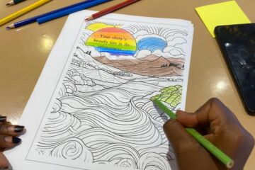 A picture of someone coloring in a print out that says Every journey needs a first step. You can see someones hands, a blue colour pen and the printout half coloured with blue, yellow, red, black and purple.