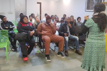 The photo is from an awareness session about legal pathways for refugees. A small group of adults are sitting on chairs and listening to a woman speak.