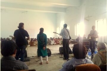 [:en]People on stage with instruments, children sitting on the floor in rows[:fi]Neljä ihmistä soittaa soittimia, kymmeniä lapsia istuu lattialla riveissä[:fr]People on stage with instruments[:]